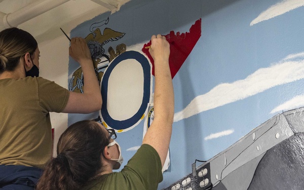 Two individuals wearing olive/green military-style shirts and face masks are painting a large wall mural. The mural depicts a naval scene including a warship hull in gray tones, a blue sky with clouds, and what appears to be a military seal or emblem with an eagle, a circular element, and red coloring. Both painters are actively working on different sections of the mural simultaneously.