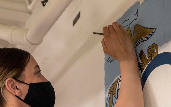 A person wearing a black face mask and olive/tan t-shirt is painting a mural on a wall inside what appears to be a military vessel or facility. The mural features patriotic and military imagery including an eagle, a large letter 'O' in blue and white, and red elements. A small patch or emblem visible in the lower right corner suggests a U.S. Navy affiliation.