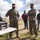 A group of military personnel in MARPAT camouflage uniforms and civilians gather outdoors around a folding table with electronic equipment. One uniformed individual appears to be briefing or demonstrating the equipment to the group. All individuals are wearing black face masks, consistent with COVID-19 era protocols.