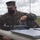 A U.S. Marine Corps officer in MARPAT camouflage uniform and black face mask is examining networking equipment on a field table in an outdoor environment. The officer appears to be inspecting or configuring a wireless router with multiple antennas alongside laptop computers. The setting appears to be a field exercise or demonstration area with open terrain visible in the background.