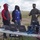 A U.S. Marine in camouflage uniform appears to be briefing or demonstrating equipment to two civilians and another service member at an outdoor field location. The group is gathered around a folding table equipped with multiple laptops, networking hardware, and cabling. All individuals are wearing face masks, suggesting this was taken during the COVID-19 pandemic period.