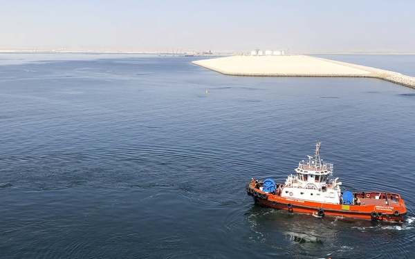 The image is taken from the flight deck of a large naval vessel, likely a US Navy amphibious assault ship, showing an orange-and-white harbor tugboat operating in calm blue waters. A military aircraft, appearing to be an AV-8B Harrier or similar jet with Marine Corps markings and a knight insignia, is visible in the foreground on the flight deck. In the background, a sandy artificial breakwater or land reclamation feature is visible, suggesting a Middle Eastern port environment.