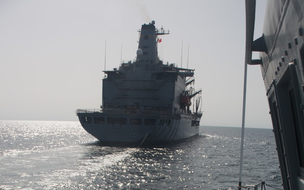 The image shows two naval vessels conducting an underway replenishment (UNREP) or connected replenishment operation at sea. A warship (foreground, right) is positioned alongside a larger auxiliary/replenishment vessel in the background. Crew members in hard hats are visible on the deck of the foreground vessel, managing lines or equipment.