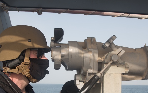 A military or naval personnel member is using a large mounted optical surveillance device (big eyes/binoculars) aboard a naval vessel at sea. The individual is wearing full tactical gear including a combat helmet, face mask, gloves, and communications equipment. The setting appears to be an open bridge wing or deck with calm ocean waters visible in the background.