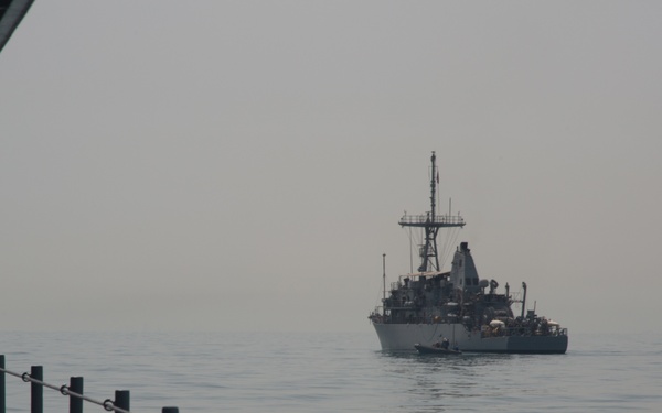 A naval warship is observed at a distance across calm, flat water under hazy, overcast conditions. The photograph is taken from the deck of another vessel, with a metal safety railing visible in the foreground. The distant ship appears to be a military vessel with a prominent mast, radar equipment, and superstructure.