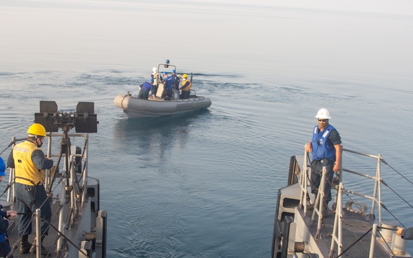 Naval personnel are conducting a small boat operations exercise from the stern of a military vessel. A rigid inflatable boat (RIB) with crew members aboard is positioned in the water just off the ship's stern ramp. Personnel on deck are wearing safety helmets and face masks while overseeing the operation.