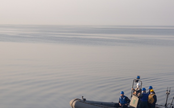 A rigid inflatable boat (RIB) with approximately four to five personnel aboard is underway on calm, glassy water under hazy atmospheric conditions. The crew members are wearing safety helmets and life vests, and an American flag is visible at the stern. The boat appears to be a military or law enforcement vessel based on its gray color scheme and equipment.