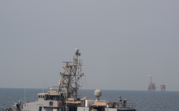 A U.S. Navy patrol coastal ship named 'SQUALL' is underway at speed on open ocean waters. The vessel is painted in standard naval gray and is equipped with various radar and communications equipment on its mast structure. In the background, an offshore oil or gas platform is visible on the horizon with a visible flare stack burning.