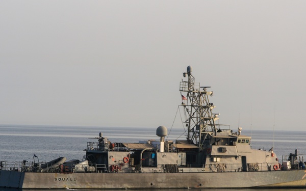 A U.S. Navy patrol coastal vessel named 'SQUALL' is photographed underway or at slow speed on calm, open water under overcast skies. The vessel displays an American flag and is equipped with various weapons systems, radar equipment, and communications masts. The image appears to be an official or documentary naval photograph taken in a maritime operational environment.