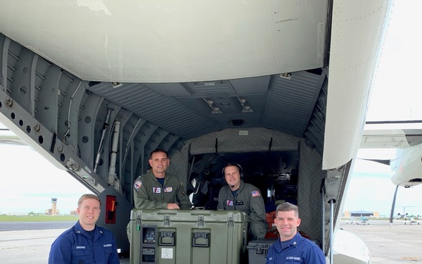 Four military personnel are posed near the open cargo ramp of a large military aircraft on an airfield tarmac. Two individuals in blue U.S. Coast Guard coveralls stand at ground level in front of the ramp, while two others in olive/green flight suits sit inside the aircraft's cargo bay. A large olive-drab military equipment case is visible on the cargo ramp.