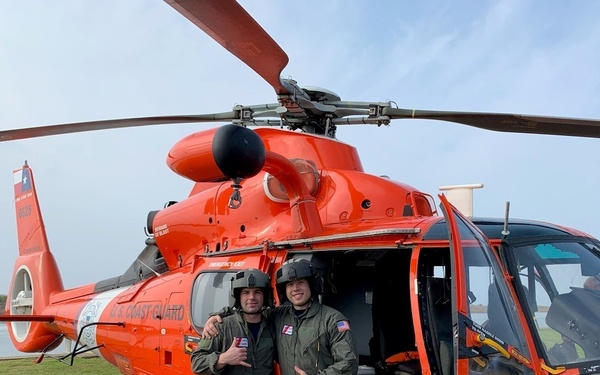 Two U.S. Coast Guard personnel in olive green flight suits and helmets pose in front of an orange MH-65 Dolphin rescue helicopter. Both individuals are giving thumbs-up gestures, suggesting a successful mission or positive occasion. The helicopter's side door is open, revealing cockpit instrumentation and additional crew inside.