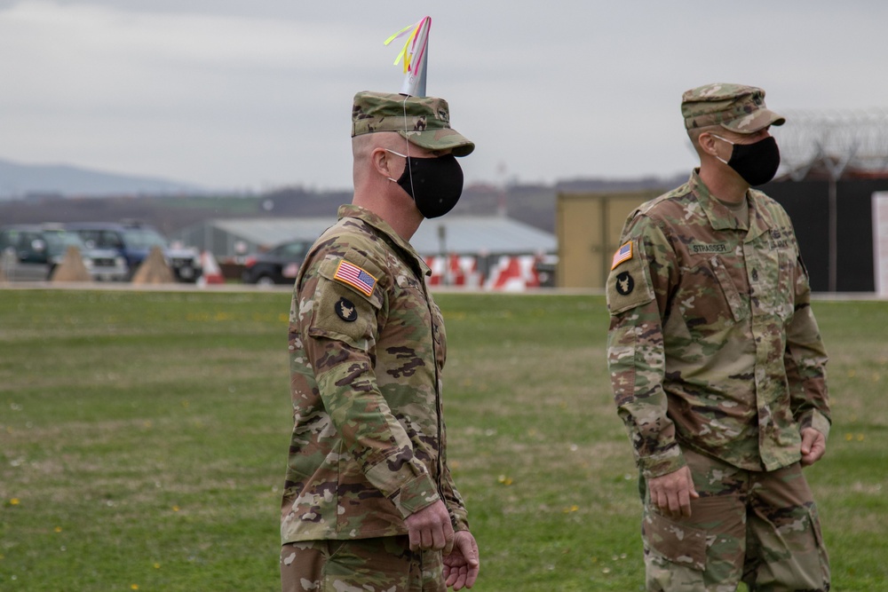 Two U.S. Army soldiers in OCP (Operational Camouflage Pattern) uniforms and black face masks are standing on a grass field, likely at a military installation. The soldier in the foreground is wearing a colorful party hat on top of his patrol cap, suggesting a celebratory occasion. A name tape reading 'STRASSER' is visible on the soldier in the background.