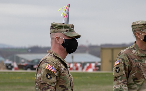 Two U.S. Army soldiers in OCP (Operational Camouflage Pattern) uniforms and black face masks are standing on a grass field, likely at a military installation. The soldier in the foreground is wearing a colorful party hat on top of his patrol cap, suggesting a celebratory occasion. A name tape reading 'STRASSER' is visible on the soldier in the background.