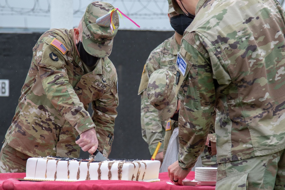 U.S. Army soldiers in OCP (Operational Camouflage Pattern) uniforms are gathered around a table with a large white decorated cake, with one soldier cutting it. The setting appears to be an outdoor or semi-outdoor military ceremony, likely a unit birthday or celebration event. All visible personnel are wearing black face masks, suggesting this was taken during the COVID-19 pandemic period.