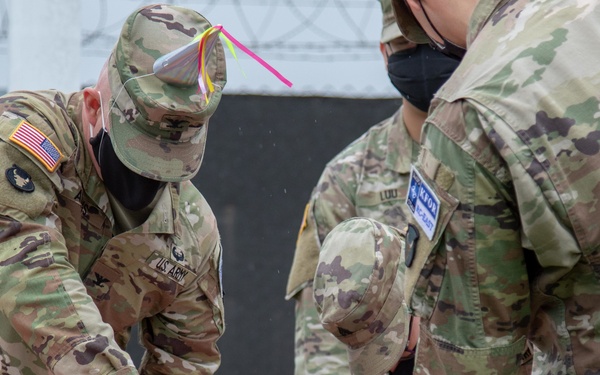 U.S. Army soldiers in OCP (Operational Camouflage Pattern) uniforms are gathered around a table with a large white decorated cake, with one soldier cutting it. The setting appears to be an outdoor or semi-outdoor military ceremony, likely a unit birthday or celebration event. All visible personnel are wearing black face masks, suggesting this was taken during the COVID-19 pandemic period.