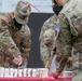 U.S. Army soldiers in OCP (Operational Camouflage Pattern) uniforms are gathered around a table with a large white decorated cake, with one soldier cutting it. The setting appears to be an outdoor or semi-outdoor military ceremony, likely a unit birthday or celebration event. All visible personnel are wearing black face masks, suggesting this was taken during the COVID-19 pandemic period.