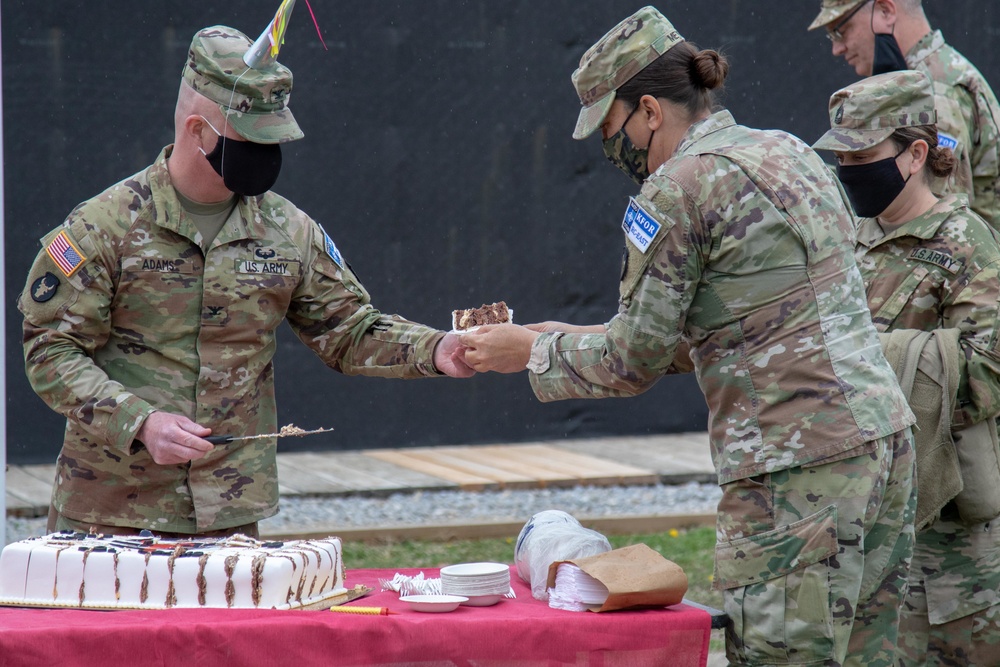 U.S. Army soldiers in OCP (Operational Camouflage Pattern) uniforms are participating in a military cake-cutting ceremony, likely a unit birthday or anniversary celebration. A soldier identified by the name tape 'ADAMS' is holding a ceremonial sword/saber used to cut the cake, while another soldier hands him a slice. All visible personnel are wearing black face masks, consistent with COVID-19 era protocols.