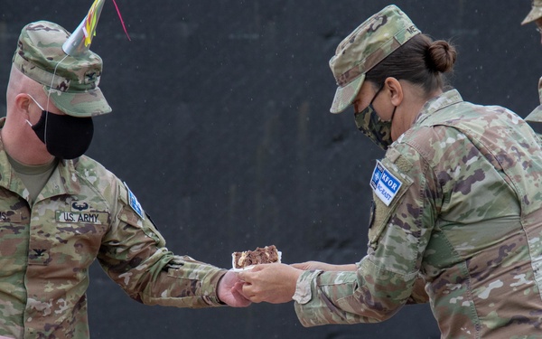 U.S. Army soldiers in OCP (Operational Camouflage Pattern) uniforms are participating in a military cake-cutting ceremony, likely a unit birthday or anniversary celebration. A soldier identified by the name tape 'ADAMS' is holding a ceremonial sword/saber used to cut the cake, while another soldier hands him a slice. All visible personnel are wearing black face masks, consistent with COVID-19 era protocols.