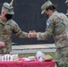 U.S. Army soldiers in OCP (Operational Camouflage Pattern) uniforms are participating in a military cake-cutting ceremony, likely a unit birthday or anniversary celebration. A soldier identified by the name tape 'ADAMS' is holding a ceremonial sword/saber used to cut the cake, while another soldier hands him a slice. All visible personnel are wearing black face masks, consistent with COVID-19 era protocols.