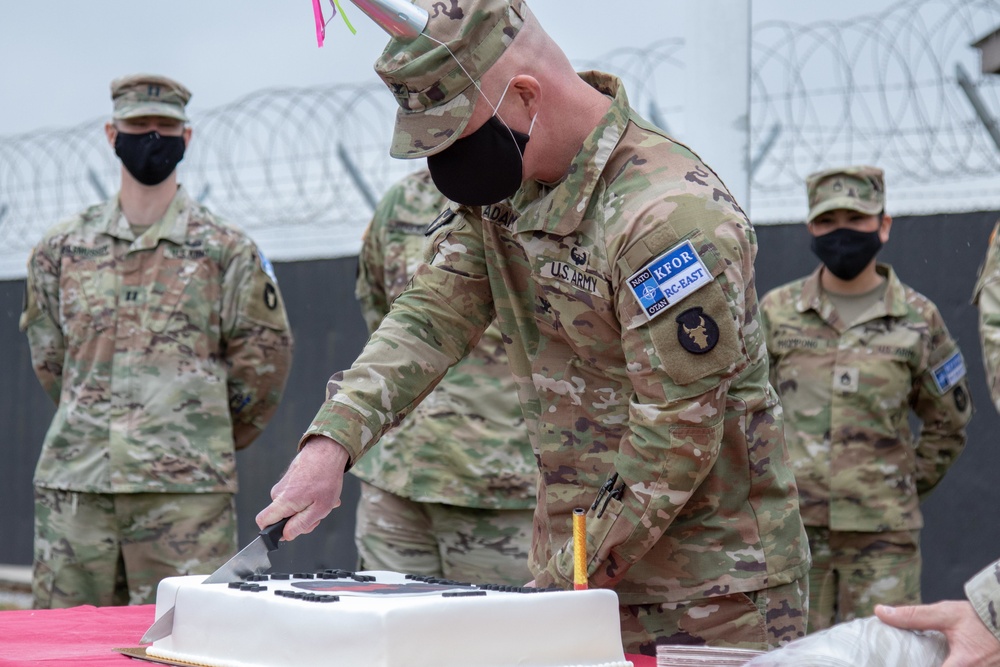 A U.S. Army soldier wearing a NATO KFOR RC-East patch is cutting a white ceremonial cake, likely during a military anniversary or celebration event. Several other uniformed soldiers wearing black face masks stand in the background near a razor wire barrier. The setting appears to be an outdoor military installation.