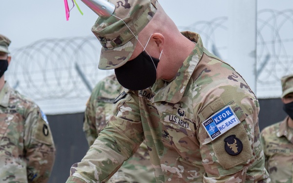 A U.S. Army soldier wearing a NATO KFOR RC-East patch is cutting a white ceremonial cake, likely during a military anniversary or celebration event. Several other uniformed soldiers wearing black face masks stand in the background near a razor wire barrier. The setting appears to be an outdoor military installation.