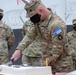 A U.S. Army soldier wearing a NATO KFOR RC-East patch is cutting a white ceremonial cake, likely during a military anniversary or celebration event. Several other uniformed soldiers wearing black face masks stand in the background near a razor wire barrier. The setting appears to be an outdoor military installation.