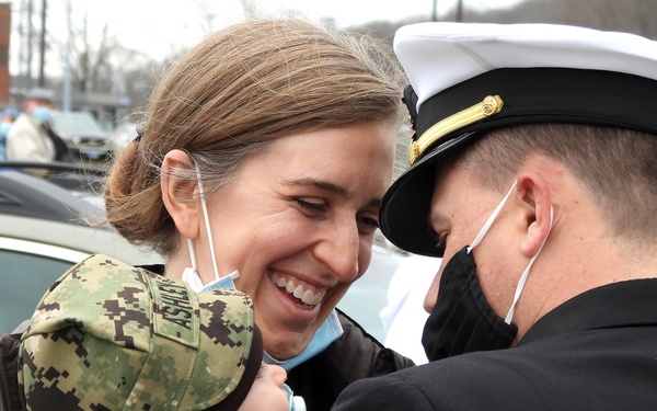 A joyful reunion scene showing a smiling woman holding an infant dressed in a miniature military camouflage uniform, greeting a person in a dark Navy dress uniform with a white officer's cover. Both adults are wearing face masks pulled down or partially worn, suggesting a COVID-era military homecoming event. The emotional moment appears to take place in a parking lot setting.