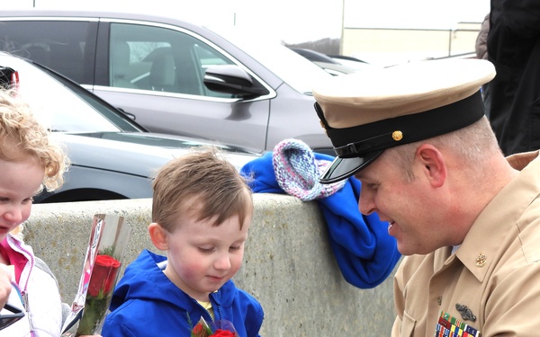 A uniformed military officer, appearing to be a U.S. Navy or Marine Corps senior enlisted member, crouches down to interact with two young children who are holding single red roses wrapped in foil. The scene appears to be a homecoming or welcome ceremony, likely at a military base, with parked vehicles visible in the background. The children, a girl with curly blonde hair and a boy in a blue jacket, appear happy and engaged with the servicemember.