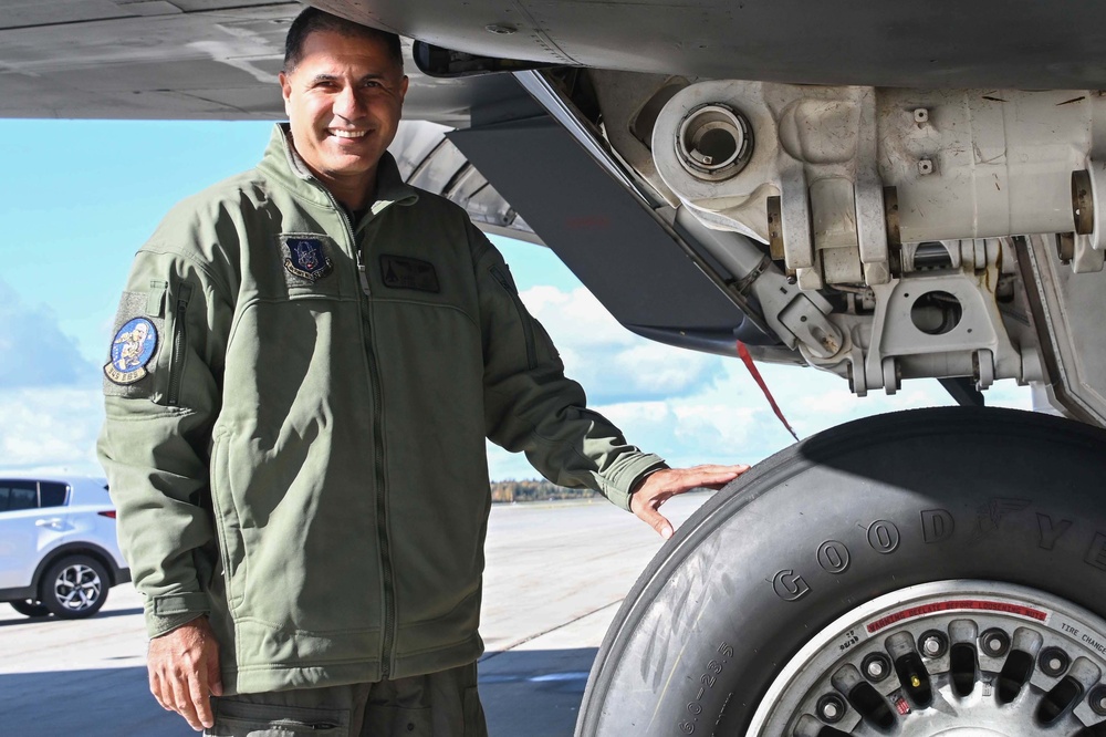 A military aviator in an olive green flight suit stands beside the landing gear of a large military aircraft on a tarmac. He is smiling and resting his hand on a Goodyear tire mounted on the aircraft's main landing gear assembly. The image appears to be an official or semi-official portrait-style photograph taken outdoors in clear weather.