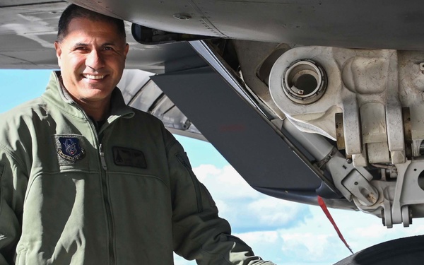 A military aviator in an olive green flight suit stands beside the landing gear of a large military aircraft on a tarmac. He is smiling and resting his hand on a Goodyear tire mounted on the aircraft's main landing gear assembly. The image appears to be an official or semi-official portrait-style photograph taken outdoors in clear weather.