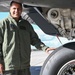 A military aviator in an olive green flight suit stands beside the landing gear of a large military aircraft on a tarmac. He is smiling and resting his hand on a Goodyear tire mounted on the aircraft's main landing gear assembly. The image appears to be an official or semi-official portrait-style photograph taken outdoors in clear weather.