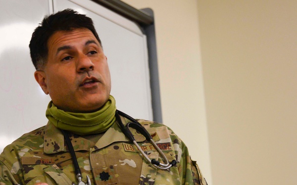 A U.S. Air Force military medical officer in OCP (Operational Camouflage Pattern) uniform is presenting or lecturing in front of a whiteboard. He is wearing a stethoscope around his neck and a green neck gaiter pulled down, holding markers in both hands. His rank insignia appears to be that of a Major (oak leaf cluster visible on chest).