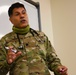 A U.S. Air Force military medical officer in OCP (Operational Camouflage Pattern) uniform is presenting or lecturing in front of a whiteboard. He is wearing a stethoscope around his neck and a green neck gaiter pulled down, holding markers in both hands. His rank insignia appears to be that of a Major (oak leaf cluster visible on chest).