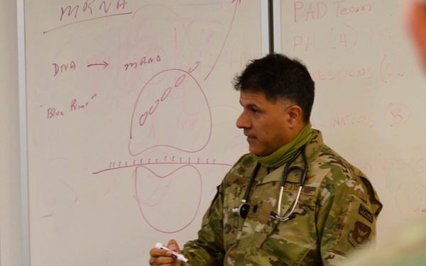 A military medical officer in camouflage uniform with a stethoscope around his neck is conducting a briefing or training session in front of a whiteboard. The whiteboard contains diagrams and text related to mRNA, DNA, and what appears to be a cell diagram, suggesting a medical or vaccine education briefing. Other uniformed personnel are visible in the foreground as the audience.