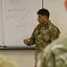 A military medical officer in camouflage uniform with a stethoscope around his neck is conducting a briefing or training session in front of a whiteboard. The whiteboard contains diagrams and text related to mRNA, DNA, and what appears to be a cell diagram, suggesting a medical or vaccine education briefing. Other uniformed personnel are visible in the foreground as the audience.