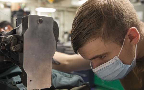 A young man wearing a light blue surgical mask and olive/brown t-shirt is operating an industrial sewing machine, closely inspecting his work. He is sewing what appears to be olive drab military-style fabric or gear. A second person wearing safety glasses is visible in the background at another workstation.