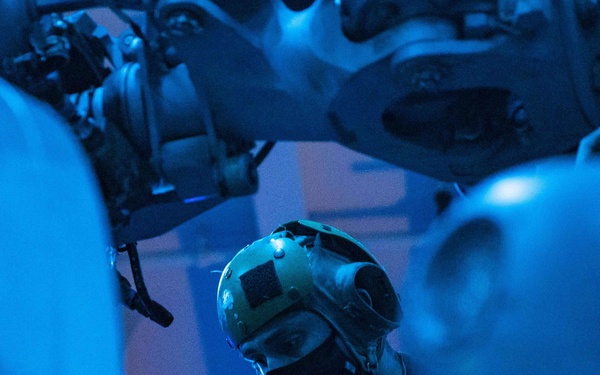 A technician or military maintenance personnel is working on mechanical or aircraft equipment in a blue-lit environment, likely a hangar deck or maintenance bay. The individual is wearing a protective helmet with a velcro patch and a black face mask while focused on the machinery in front of them. The scene is bathed in deep blue lighting, suggesting a low-light or night operations environment.