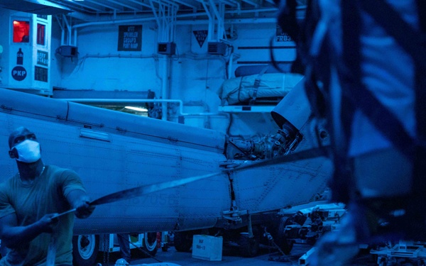 A naval personnel member wearing a face mask and eye protection works in what appears to be an aircraft carrier hangar bay under blue lighting conditions. The scene shows maintenance or ordnance handling activities near a military aircraft, with various equipment and support vehicles visible in the background. The dominant blue lighting suggests nighttime operations or a specific lighting protocol in use aboard the vessel.