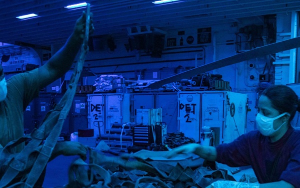 Two military personnel wearing face masks and gloves are handling a large cargo bundle secured with netting and straps in what appears to be a ship's hangar bay. The space is illuminated with a distinctive blue light, revealing helicopter components and equipment crates marked 'DET 2' in the background. The scene suggests a logistics or cargo handling operation aboard a naval vessel.