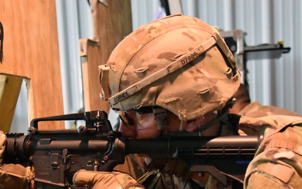 A soldier in MultiCam/OCP camouflage uniform is aiming an M4/M16-style rifle through a gap in a plywood barrier marked with the number '9'. The soldier is wearing a tactical helmet with visible markings and is in a shooting stance during what appears to be a training exercise. A unit patch resembling the U.S. Army 'A' patch is visible on the right sleeve.