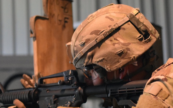 A soldier in full multicam camouflage uniform and helmet is aiming an M16/M4-style rifle from behind a wooden barrier or barricade. The soldier appears to be in a training or range environment, indicated by the indoor/covered facility setting. Tactical gear including gloves, helmet cover, and unit patches are clearly visible.