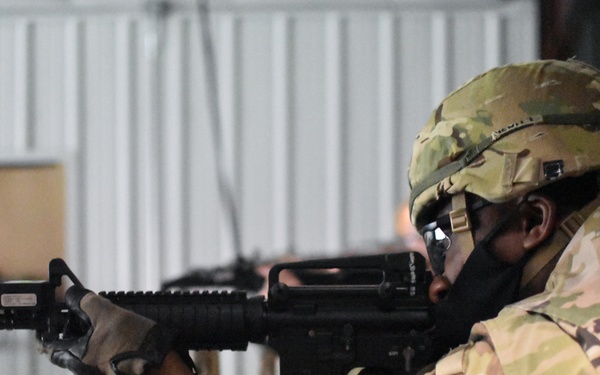 A soldier in full camouflage uniform and tactical helmet is aiming an M4/M16-style rifle at a shooting range. The individual is wearing OCP (Operational Camouflage Pattern) gear, protective eyewear, and a face covering. The setting appears to be an indoor or covered firing range with target silhouettes visible in the background.
