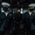 Three U.S. Navy personnel are stationed on the bridge of a naval vessel, all wearing dark face masks. Two sailors in enlisted dress blue uniforms with white Dixie cup covers flank a petty officer in a service dress blue uniform wearing a headset. They appear to be on watch duty at a navigation or helm console.
