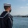 A U.S. Navy sailor in dress blues uniform stands at the rail of a naval vessel, looking out toward a city skyline across a body of water. The sailor is wearing a black face mask, a white dixie cup hat, and a uniform with a ship name tag visible on the shoulder reading 'USS JOHN FINN'. The background shows a coastal city skyline, likely San Diego, California, under a clear blue sky.
