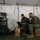 Marines with 3d Supply Battalion demonstrate their field communication capabilities Two military personnel in camouflage uniforms and face masks are working on laptops inside a temporary field tent. They are seated on cardboard boxes next to a rack of networking and server equipment. The setup appears to be a mobile tactical operations or communications center.