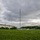 Marines with 3d Supply Battalion demonstrate their field communication capabilities A person in military/camouflage clothing is setting up or adjusting a field antenna system on a grassy open area. The antenna consists of a central vertical mast with multiple wire elements extending outward in a fan or inverted-V configuration, consistent with a military HF radio antenna. The setting appears to be a military installation with institutional buildings and power lines visible in the background.