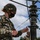 Marines with 3d Supply Battalion demonstrate their field communication capabilities A U.S. Marine in MARPAT digital camouflage uniform and black face mask is working on what appears to be a military antenna or communications mast assembly. The service member is handling cables and rigging components attached to a vertical pole with multiple disc-shaped brackets. A second uniformed individual is partially visible in the background.