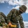 Marines with 3d Supply Battalion demonstrate their field communication capabilities A U.S. Marine in MARPAT digital camouflage uniform is kneeling on a gravel surface, using a sledgehammer with a yellow handle to drive a metal stake or spike into the ground. The Marine is wearing a tactical face mask, glasses, a Kevlar helmet, and tan combat boots. The name tape reads 'U.S. MARINES' and a partial surname is visible on the chest.