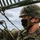 Marines with 3d Supply Battalion demonstrate their field communication capabilities A U.S. Marine in MARPAT digital camouflage uniform and helmet is working on what appears to be a military antenna or communications equipment. The service member is wearing a black face mask and tan helmet chin strap, suggesting this image was taken during the COVID-19 pandemic era. The photo is a close-up shot with a partially cloudy sky visible in the background.