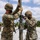 Marines with 3d Supply Battalion demonstrate their field communication capabilities Two military personnel in MARPAT digital camouflage uniforms and combat helmets are assembling or adjusting a communications mast or antenna pole in an outdoor gravel area. Both individuals are wearing black face masks and tactical gloves while working on the mast's rigging hardware and guy-wire attachments. A third service member and a green military tent are visible in the background.